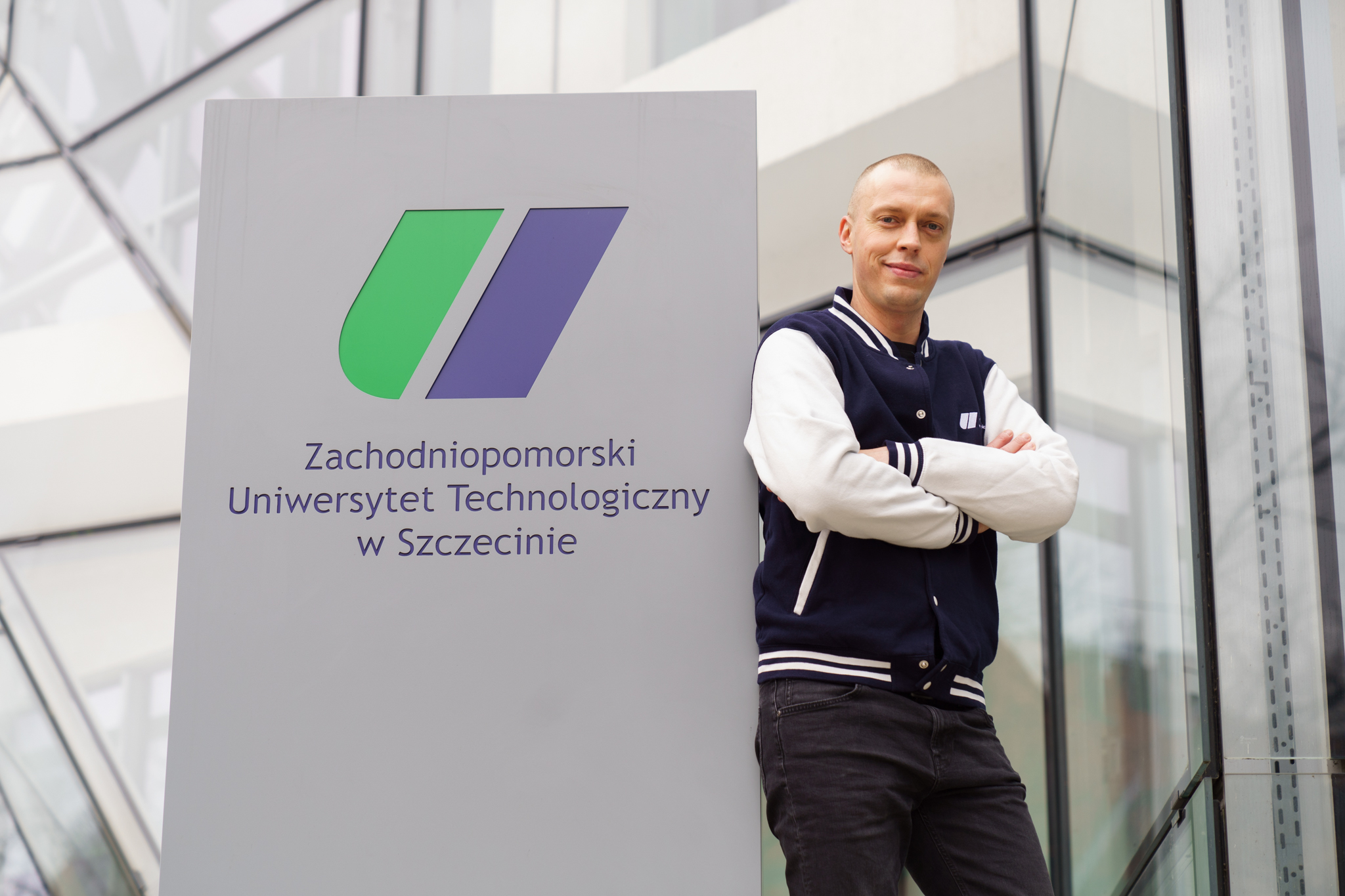 Osoba stoi obok tablicy z napisem 'Zachodniopomorski Uniwersytet Technologiczny w Szczecinie' oraz logo składającym się z zielonych i niebieskich kształtów. Osoba ma założoną granatowo-białą kurtkę i skrzyżowane ramiona.